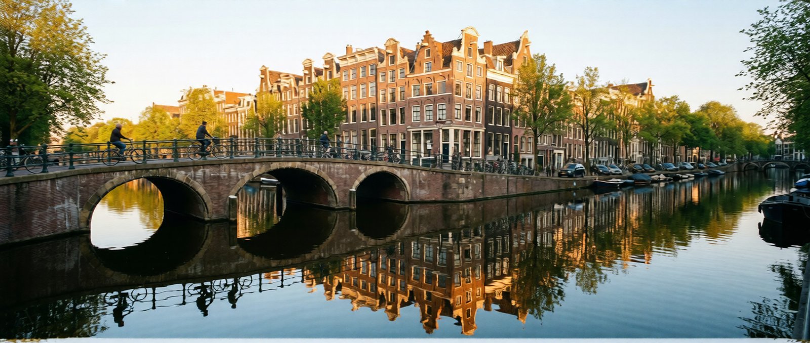 A scenic view of Amsterdam's canals with historic houses and a cyclist on a bridge during sunset.